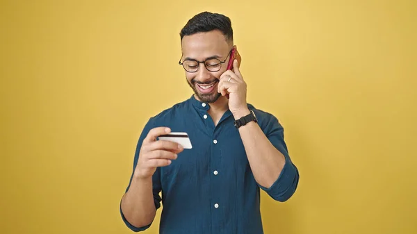 Young hispanic man speaking on the phone holding credit card over isolated yellow background