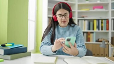 Genç İspanyol kız öğrenci akıllı telefon kullanıyor kulaklık takıyor kütüphane üniversitesinde notlar alıyor.
