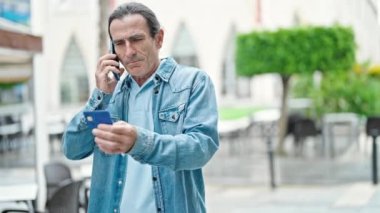 Orta yaşlı bir adam elinde kredi kartıyla kahve dükkanının terasında akıllı telefonla tartışıyor.