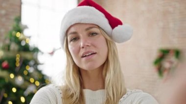 Genç sarışın kadın, evde Noel ağacının yanında el sallayarak video çekiyor.