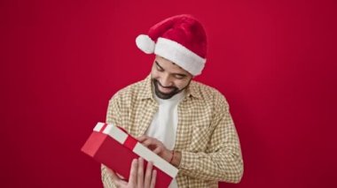 Genç İspanyol adam, kendine güvenen, Noel şapkası takan, hediye duyan, izole edilmiş kırmızı arka plan üzerinde ses çıkaran bir adam.