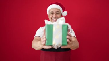 Gri saçlı orta yaşlı bir adam izole edilmiş kırmızı arka planda noel hediyesi tutuyor.