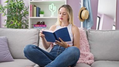 Young blonde woman reading book sitting on sofa at home