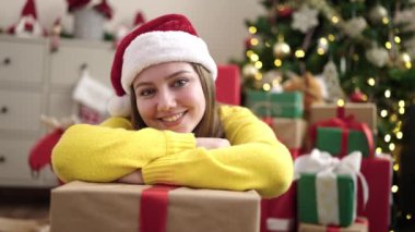 Hediyeye yaslanan genç sarışın kadın evdeki Noel ağacının yanında yerde oturuyor.