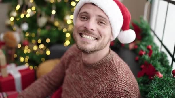 Νεαρός Καυκάσιος Κάθεται Στον Καναπέ Δίπλα Στο Χριστουγεννιάτικο Δέντρο Φυσώντας — Αρχείο Βίντεο