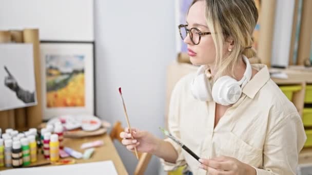 在艺术工作室里 年轻的金发女艺术家坐在桌子上选择带耳机的画笔 — 图库视频影像