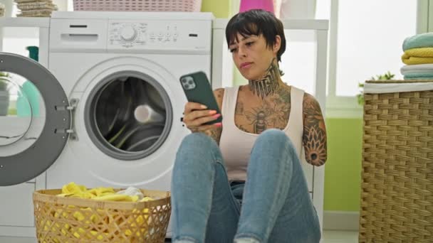 西班牙裔妇女 断臂时使用智能手机 坐在洗衣房洗衣机旁 — 图库视频影像