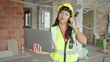 Genç Çinli kadın mimar inşaat alanında bilgisayarla akıllı telefondan konuşuyor.