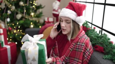 Genç sarışın kadın akıllı telefonuyla konuşuyor evdeki noel ağacının yanında koltukta oturuyor.