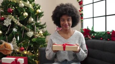 Afrikalı Amerikalı kadın elinde hediyeyle evdeki Noel ağacının yanındaki koltukta oturuyor.