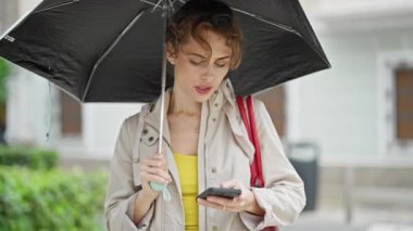 Sokakta gülümseyen akıllı telefon kullanan şemsiyeli genç bir kadın.
