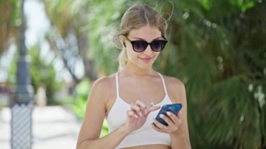 Genç sarışın kadın parkta akıllı telefondan müzik dinliyor.