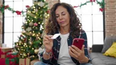 Orta yaşlı İspanyol kadın akıllı telefon ve kredi kartıyla Noel 'i evde kutluyor.