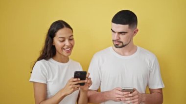 Güzel bir ana yakalanmış sevimli bir çift, akıllı telefonlara yazarken özgüvenle gülümsüyorlar, izole edilmiş sarı bir arka plana karşı.
