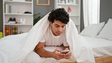 Genç Latin adam yatak odasına sürpriz bir şekilde battaniyeli akıllı telefon kullanıyor.