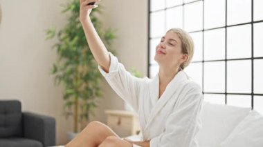 Bornoz giymiş genç sarışın kadın yatakta oturmuş yatak odasında akıllı telefondan selfie çekiyor.