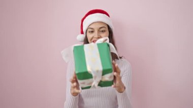 Noel şapkası takmış gülümseyen genç, güzel İspanyol kadın izole edilmiş pembe arka plandan hediyeyi dinliyor.