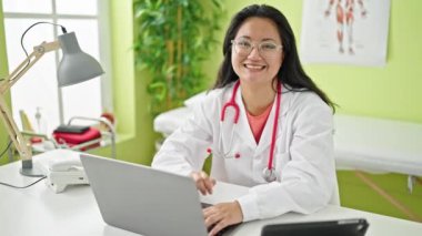Genç Çinli doktor dizüstü bilgisayar kullanıyor. Klinikte kollarını kavuşturup oturuyor.