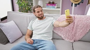 Evdeki kanepede oturan ve akıllı telefonuyla selfie çeken İspanyol genç adam.