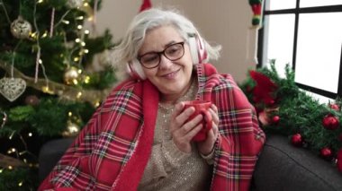 Orta yaşlı, kır saçlı, müzik dinleyen, kahve içen, Noel ağacının yanında oturan bir kadın.