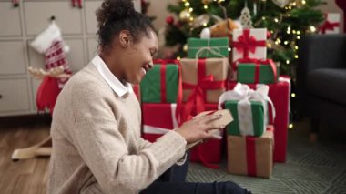 Afrikalı Amerikalı bir kadın elinde hediyeyle evde Noel ağacının yanında oturuyor.