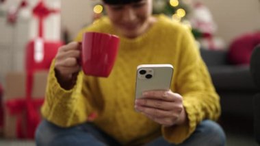 Genç Çinli kadın akıllı telefon kullanıyor. Evde Noel ağacının yanında kahve içiyor.