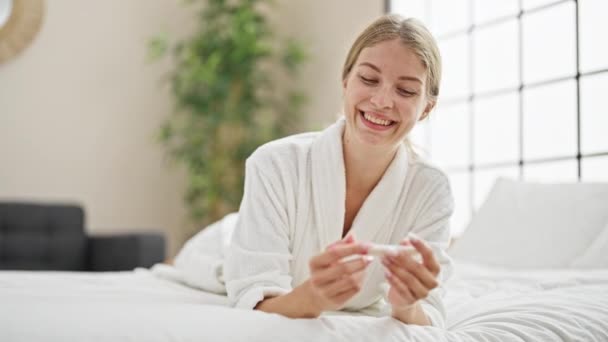 ベッドルームで妊娠テストを保持するバスローブを着用している若い金髪の女性 — ストック動画