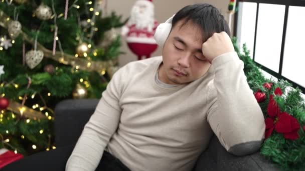Молодой Китаец Слушает Музыку Спящую Диване Рождественской Елки Дома — стоковое видео