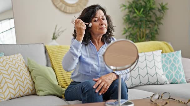 中年ヒスパニック系女性が自宅で鏡を見ながらソファーに座って顔をマッサージ — ストック動画