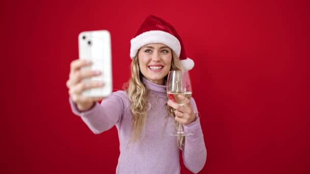 クリスマスの帽子をかぶった若い金髪の女性は 隔離された赤い背景の上にワインのグラスを保持するビデオコールを持っています — ストック動画