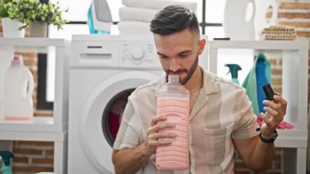 クリーンタオルと洗剤瓶の匂いを嗅ぐ若いヒスパニック男性 洗濯室で笑顔 — ストック動画