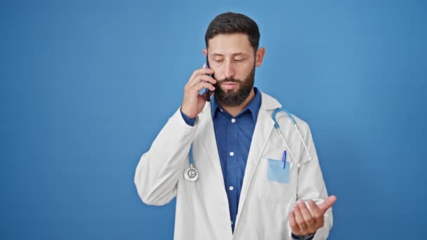 孤立した青い背景を見ているスマートフォンで話す若いヒスパニック男性の医師 — ストック動画