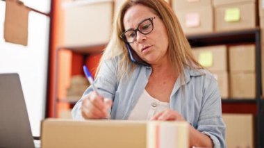 Orta yaşlı İspanyol kadın ecommerce iş işçisi pakete yazı yazıp ofisteki akıllı telefondan konuşuyor.