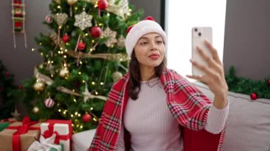 Genç ve güzel İspanyol kadın evdeki Noel ağacının yanında kanepeye oturmuş video görüşmesi yapıyor.