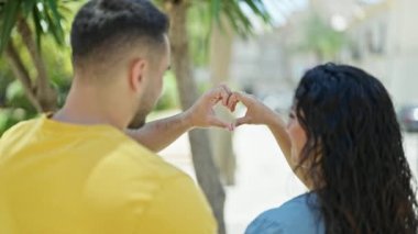 Kadın ve erkek, parkta kalp hareketi yaparken gülümsüyor.