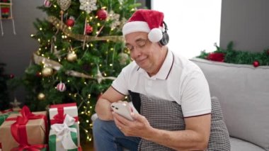 Gri saçlı orta yaşlı bir adam telefon kullanıyor. Evde Noel süslemeleri yaparak müzik dinliyor.