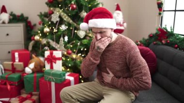 Genç kel adam yılbaşı ağacının yanında oturmuş evde karın ağrısı çekiyor.