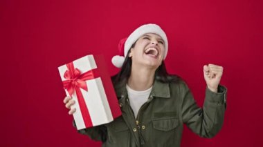 Genç, güzel, İspanyol bir kadın elinde Noel hediyesi tutarak izole edilmiş kırmızı arka planda dans ediyor.