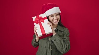 Genç, güzel, İspanyol bir kadın, gülümsüyor. Kendine güveniyor, kırmızı arka planda bir Noel hediyesi tutuyor.