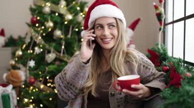 Genç sarışın kadın akıllı telefonda konuşuyor. Evdeki Noel ağacının yanında kanepede kahve içiyor.
