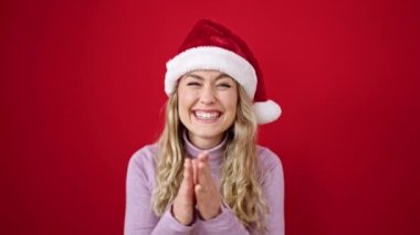 Genç sarışın kadın, Noel şapkası takıyor, el çırpıyor, kırmızı arka planda alkışlıyor.