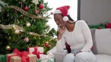 Afrikalı Amerikalı kadın elinde hediyeyle evdeki Noel ağacının yanındaki koltukta oturuyor.