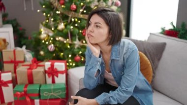 Kafkasyalı genç bir kadın Noel ağacının yanında kanepeye oturmuş üzgün üzgün bakıyor.