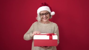 Gri saçlı orta yaşlı bir kadın Noel şapkası takıyor ve izole edilmiş kırmızı arka planda hediyesini açıyor.