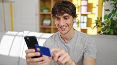 Genç İspanyol adam akıllı telefon ve kredi kartıyla alışveriş yapıyor. Evdeki kanepede oturuyor.