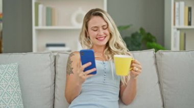 Genç sarışın kadın akıllı telefon kullanıyor. Evde kahve içiyor.