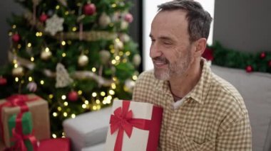 Orta yaşlı bir adam elinde hediyeyle evde Noel ağacının yanında oturuyor.