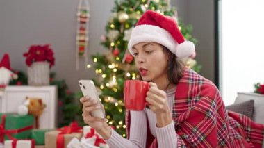 Genç ve güzel İspanyol kadın akıllı telefon kullanıyor Noel ağacının yanında oturmuş evde kahve içiyor.