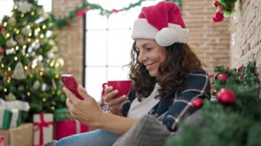 Orta yaşlı İspanyol kadın akıllı telefon kullanıyor evde Noel 'i kutlamak için kahve içiyor.