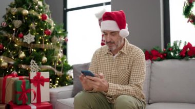 Orta yaşlı bir adam evdeki Noel ağacının yanındaki koltukta akıllı telefon kullanıyor.